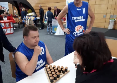 Teilnehmer der TSV 1880 Schwandorf Tigers spielt ein Brettspiel bei der Inklu‑Con in der Stadthalle Burglengenfeld, umgeben von Besucherinnen und Besuchern